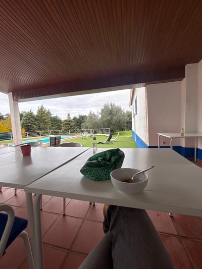 The image depicts a covered outdoor patio area in Montemor-o-Novo, Portugal, overlooking a swimming pool and garden under an overcast sky. In the foreground, a person's legs, clad in grey jeans and brown shoes, are visible beneath a white rectangular table. On the table, from left to right, are a pink cylindrical pot, a green patterned cloth bag, a clear glass bottle with a silver cap, and a white bowl containing a spoon and a light-colored liquid. The patio floor is covered with reddish-brown tiles. The patio ceiling is constructed from dark brown slatted panels. Beyond the patio, a blue-tiled swimming pool is visible within a green lawn area. The lawn features several trees, including olive trees with silvery-green foliage and darker evergreen trees. White lounge chairs and small white tables are scattered across the grass. To the right, a white building with a distinctive blue stripe along its base is visible, and two additional white tables are situated near its exterior wall, each holding a small yellow-capped bottle.
