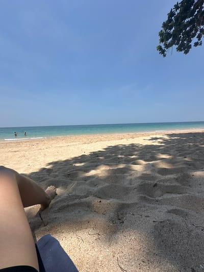 This image depicts a sunny beach scene in Phra Ae, Thailand. The foreground shows the lower legs and bare feet of an individual resting on light-colored sand, with a dark blue, possibly ribbed, fabric item positioned beneath the legs. Dappled shadows from an overhead tree extensively cover the sandy surface, which displays subtle ripples and scattered organic debris. In the midground, the calm, clear turquoise ocean extends to the horizon under a bright blue sky. Two individuals are partially submerged in the water near the shoreline, appearing to be wading or standing. Green foliage from a tree is visible in the upper right corner, contributing to the cast shadows.