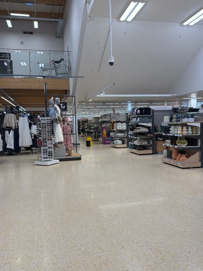 The image shows the interior of a supermarket or department store in the city of Pity Me, United Kingdom. The store has high ceilings and bright lighting from multiple fluorescent fixtures. A security camera is visible. Aisles are lined with shelving units displaying a variety of merchandise, including clothing, accessories, and home goods. Mannequins are dressed in women's clothing. Shopping carts are stacked in the background. The floor is tiled in a speckled pattern. No people are visible in the frame.