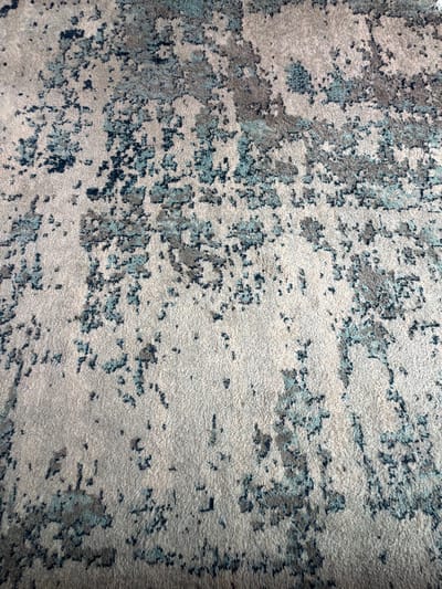 The image displays a close-up, overhead view of a textured carpet with an abstract, distressed pattern. The carpet features a base color of light gray or beige, overlaid with irregular patches and flecks of darker gray, muted teal, and deep navy blue. The pattern resembles splatters, washes of color, or the erosion of a surface, creating a mottled and variegated effect. The texture of the carpet appears soft and somewhat plush, with visible fibers contributing to the tactile quality. The lighting in the image creates subtle shadows and highlights, emphasizing the carpet's texture and depth. There is no discernible text, people, or specific scene depicted. The focus is solely on the surface of the carpet itself and its intricate design. Given the context of Dorsten, Germany, this is likely an interior furnishing.