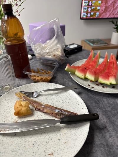 The image depicts a table setting with various food items and objects, suggestive of a meal or snack. In the foreground, a white plate with a speckled pattern holds a small piece of bread, a fork, and a partially eaten piece of fish. A knife with a black handle rests on the plate. To the left, a clear glass with vertical ridges is partially visible, and a brown plastic bottle containing a dark liquid, possibly a beverage, stands behind it.
In the middle ground, a clear plastic container holds thin, crispy snacks. Behind this, a translucent plastic bag contains white material, possibly food items. A light purple rectangular object is also visible in the background.
The background features a television screen displaying a video game interface with player statistics and avatars. To the right, a wooden stand supports a book and a small potted plant. The scene appears to be indoors, with a dark grey textured tablecloth covering the surface. There is no discernible indication of the city of Rassokha, Russia, within the provided image.