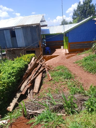 The scene depicts a rural or semi-rural setting in Kiangochi, Kenya, under a clear blue sky with scattered white clouds. The foreground consists of red-brown earth and patches of green grass and weeds. To the left, a large pile of assorted wooden planks and logs rests on the ground, adjacent to dense green shrubbery.
In the mid-ground, two primary structures are visible. The building on the left is a multi-story dwelling with a lower section constructed from grey concrete blocks and an upper section clad in corrugated metal sheeting, featuring an open, unglazed window. A pair of light-colored sandals are placed on a wooden support beam beneath the upper story. A thin, light-colored wire or string extends from this building towards the center.
To the right, a single-story building is painted with an upper blue section, an orange horizontal stripe, and a lower section with grey horizontal lines. It has a green door. Both buildings are topped with corrugated metal roofs. A covered walkway or open-air common area connects or is situated between these two structures. Within this area, a blue plastic chair is positioned, and a faint human figure appears to be seated in it. A white bucket rests near the green door of the right building.
A tall, slender pole, possibly a utility pole or antenna mast, stands upright in the center of the image. The background features lush green trees and distant hills under the same blue sky. The ground leading up to the buildings is an unpaved red-brown dirt path.