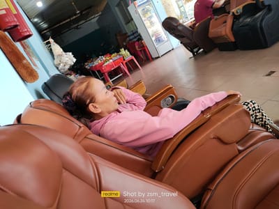 The image depicts a woman relaxing in a brown massage chair in what appears to be a commercial establishment. She is wearing a pink long-sleeved top and patterned leggings. The environment suggests a public space, possibly a salon or waiting area, with other massage chairs visible in the background. A refrigerator stocked with beverages is also present. The overall lighting suggests an indoor setting. The watermark indicates the photo was taken in Gò Dầu, Vietnam, on June 24, 2026, at 10:17 AM.