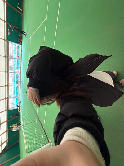 The image depicts an indoor sports facility in Souffelweyersheim, France. The primary subject, a person with long, dark hair, occupies the foreground, facing away from the camera and looking towards the distant end of a court. This individual is clad in a black long-sleeved top, an open black jacket, and visible white clothing underneath. Their right arm is raised with the hand positioned near their head. A pale-skinned forearm is visible at the bottom right, angled towards the camera. The setting features a green court marked with white lines, across which a net is stretched horizontally. In the background, beyond the net, multiple individuals are present on the court. One person, dressed in black, holds a racket near the far wall. Closer to the net, a group of approximately three to four individuals, some in yellow or light-colored attire, are visible. The facility's left side features large windows, through which residential buildings with light-colored facades and reddish roofs, along with some foliage, are visible in the exterior environment.