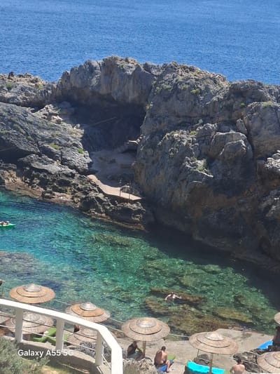 The image captures a cove along the rocky coastline in Plakias, Greece, with turquoise water. A large rock formation dominates the left side, featuring a cave-like opening accessible via a short concrete path. The right side of the frame shows a concrete platform with several straw umbrellas. People are scattered around the platform and some are swimming in the water. A white railing borders the platform.