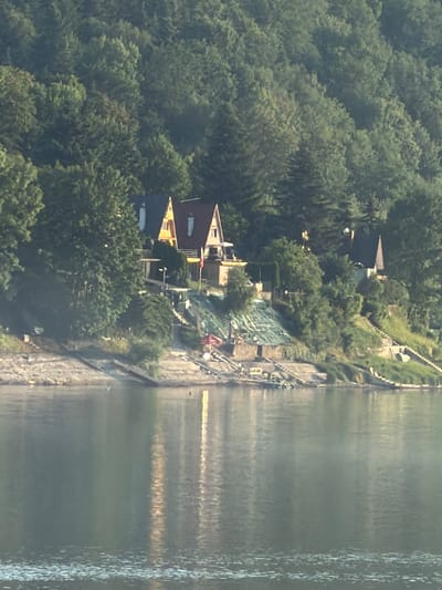 The image depicts a lakeside scene with houses built on a steep, wooded hillside. The water in the foreground reflects the trees and houses. The houses appear to be of European style, with steep, dark roofs and light-colored walls. One house in the center has a prominent yellow section. The hillside is densely covered with a variety of trees, primarily deciduous. There are pathways and structures leading up the hillside towards the houses, including stairs and what appear to be terraced areas. A red umbrella is visible on the right side of the frame, near a set of stairs. Boats or small vessels are docked or moored near the water's edge on the left side of the image. The lighting suggests it is daytime, possibly late afternoon, with some sunlight filtering through the trees and reflecting on the water. The image is of a residential area situated in a natural, possibly mountainous or hilly, environment.