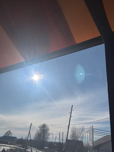 The image is an upward-angled view towards a clear blue sky with the sun and a lens flare visible. The top-left corner shows part of a building structure with orange ridged siding and a black trim. Power lines stretch across the sky, leading to a distant residential area in what could be Gory, Russia. Bare trees are visible in the background, along with rooftops and patches of snow on the ground. The buildings and objects in the residential area are unclear and somewhat obscured by distance and lighting conditions.