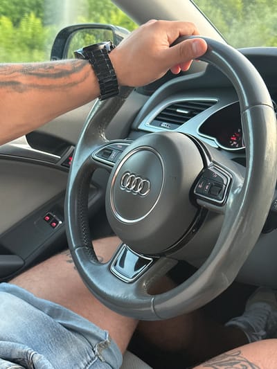 The image shows a close-up view of a person's hands on the steering wheel of a car, the dashboard, and part of the car's interior. The steering wheel is dark gray, with the Audi logo prominently displayed in the center. There are buttons on the steering wheel for various functions. The driver's left arm is visible on the left side of the steering wheel, and it has tattoos. A black watch is worn on the driver's left wrist. The driver is wearing light blue denim shorts. The car's interior appears to be gray and black. The background shows a blurred view of green foliage, suggesting the car is in motion outdoors. There is no indication of the city of Zvanka, Russia, or any specific urban environment.