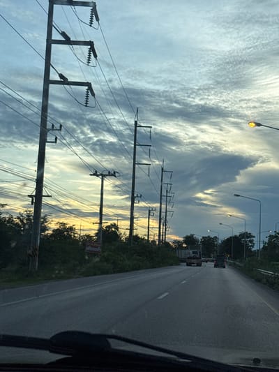 The image depicts a road in Ao Noi, Thailand, at dusk. Several utility poles with power lines are visible along the side of the road, stretching into the distance. The sky is filled with dramatic, cloudy formations, with hints of sunset colors near the horizon. Streetlights are illuminated, casting a warm glow. A few vehicles, including trucks and cars, are traveling on the road. Lush green vegetation lines the roadside. A red sign with Thai script is attached to a utility pole on the left side of the road. The foreground shows the dashboard and windshield of a vehicle, suggesting the photo was taken from inside a car.