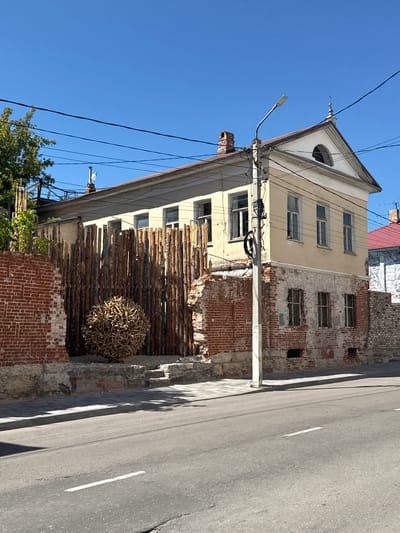 This is a full-frame, eye-level, outdoor, daytime shot of an old building on a street in Yelets, Russia. The building is two stories tall, with a light yellow facade on the upper level and exposed brick on the lower level, showing signs of decay. The building has multiple windows with white frames. A tall wooden fence, made of vertical logs, is positioned in front of the building. A large, spherical sculpture made of sticks or branches sits in front of the fence. A brick wall, partially crumbled, is to the left of the fence. A street with a white lane marker runs across the foreground, with shadows cast on it from the fence and building. A lamppost stands near the building, with power lines visible against the clear blue sky. A tree with green foliage is to the far left.