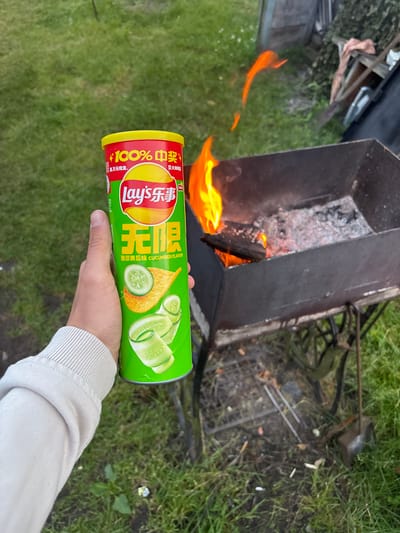 In Ганчарскі сельскі Савет, Belarus, a person's hand, wearing a cream-colored sweatshirt, holds a green can of Lay's cucumber-flavored potato chips. The can features Chinese text and imagery of cucumber slices and a potato chip. In the background, a metal barbecue grill contains burning wood and embers, with flames flickering. The scene is set outdoors on a grassy area, with the natural lighting suggesting it is either dawn or dusk. The overall impression is that of an informal outdoor gathering or a moment of relaxation.