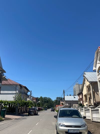 The image depicts a street scene in the city of Chahor, Ukraine. A silver sedan is parked on the right side of the road, facing away from the viewer. Several houses with various architectural styles line both sides of the street. The sky is clear and blue. The road is paved and appears to be a residential street. Power lines are visible overhead. A green trash can is located on the left side of the road near a brick fence.