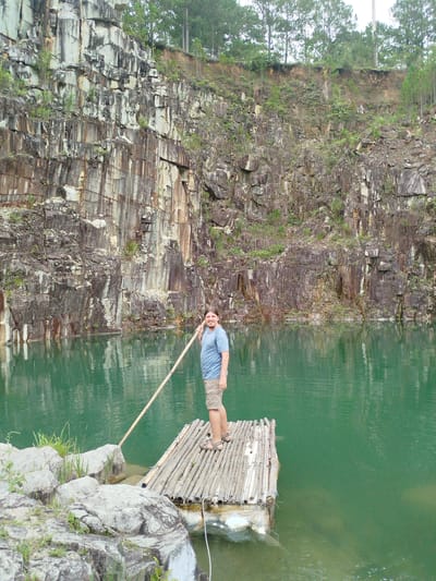 A man stands on a raft made of bamboo logs in a turquoise quarry lake. He is holding a long pole that reaches out of frame to his right. He is wearing a blue t-shirt and khaki shorts. The quarry walls are steep and made of layered rock, with some vegetation growing on them. The water is calm and reflects the sky and the quarry walls. The location is specified as the city of Lán Tranh, Vietnam.