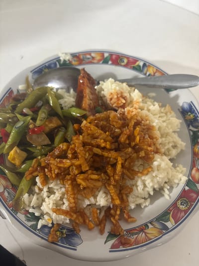 The image shows a plate of Indonesian food. The plate is white with a colorful floral rim. On the plate, there is a serving of white rice, topped with stir-fried tempeh that is cut into small strips and coated in a dark, savory sauce. To the side of the rice and tempeh is a generous portion of stir-fried green beans, mixed with pieces of tofu and red chili peppers. A spoon and a fork are resting on the plate, suggesting that the meal is ready to be eaten or is in the process of being consumed. The lighting suggests it is daytime. The food appears to be a home-cooked meal or a casual dining experience. The context provided states the location is Cibinong, Indonesia, which is consistent with the style of food. There is no visible text in the image.