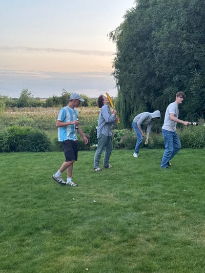 This image depicts four young adult males on a mown grass lawn in Bubwith, United Kingdom, at dusk. The background features open fields, scattered shrubbery, and a large, dense tree to the right, all beneath a pale, cloud-streaked sky. From left to right: A male wearing a blue and white striped polo shirt and a light grey baseball cap walks while holding a brown bottle. Adjacent, a male in a light blue button-up shirt holds a yellow cricket bat-like object elevated towards his head. A third male, wearing a grey hooded sweatshirt with the hood up, is bent forward, looking downwards, with a small light green ball near his left foot. The fourth male, in a light grey t-shirt and blue jeans, holds a dark, cylindrical object horizontally in his right hand, extended forward. All subjects are casually dressed and appear to be engaged in an informal outdoor activity.
