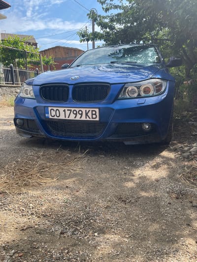 A blue BMW sedan is parked on a dirt and gravel road in Popintsi, Bulgaria. The car faces the camera and its front grille, headlights, and license plate are visible. The license plate reads "CO 1799 KB" and has a "BG" sticker. Behind the car, a tree with green leaves casts shadows on the road. In the background, a fence, a house with a vine-covered trellis, and a wooden structure are visible. The sky is blue with some clouds.