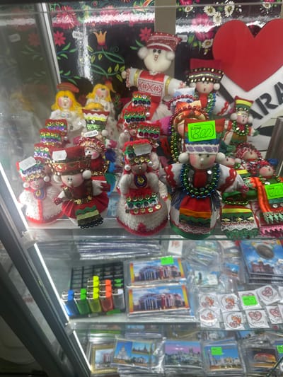 The image displays a retail display case filled with handmade dolls and various souvenir items. The dolls, predominantly female, are dressed in colorful, traditional attire adorned with beads, sequins, and embroidered details. Some dolls have price tags indicating "Цена" (price) followed by numerical values in Russian rubles, such as "800" and "600." Behind the dolls, a backdrop features a floral pattern with red and pink flowers and green leaves, interspersed with lighter flowers. A large red heart shape is visible on the right side of the backdrop, with text that appears to be a city name. Below the dolls, the display includes rows of lighters in various colors (blue, green, yellow, orange, red, grey) arranged in a rack. Further down, the case contains packaged souvenir items, including postcards or small prints depicting buildings and landmarks, and round stickers featuring cartoon foxes and heart designs. The overall scene suggests a shop selling traditional crafts and local souvenirs, possibly in Lyambir', Russia, given the context.