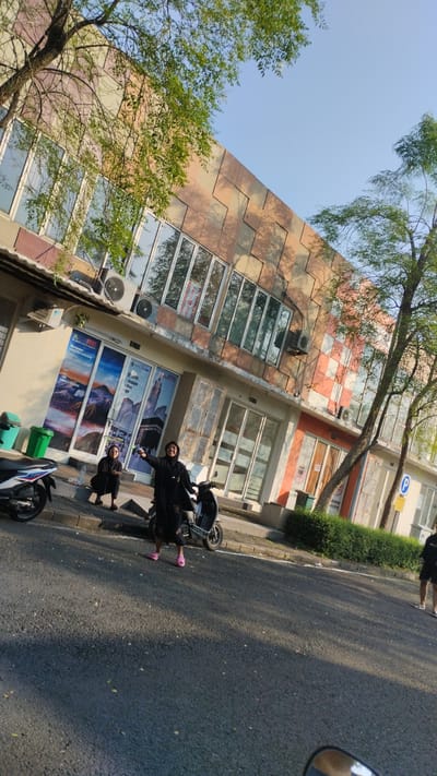 Two people are standing outside a building with a checkered facade in Trosobo, Indonesia. The building has large windows and air conditioning units on the exterior. One person is on a scooter, and the other is standing beside them. Another scooter is parked to the left. The ground is asphalt, and there are trees and bushes around the building. The sky is clear and blue.