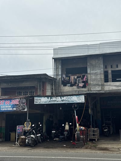 The image captures a street scene in Ajuen, Indonesia, with a focus on a collection of businesses lining the road. In the foreground, a paved road is visible, with a sidewalk or roadside area to its right. Several motorcycles are parked along the front of the buildings, suggesting a commercial area that caters to vehicle services or sales.
The buildings themselves appear to be multi-story structures with a mix of architectural styles. One building on the left features a corrugated metal roof and a banner with text, partially obscured, reading "AT 99 JOK - ALAS DASAR - PLAFON MOBIL, DLL". Next to it, a larger building displays a prominent sign for "R&J JAYA MOTOR", indicating a motorcycle repair shop or dealership. This sign also lists services like "MELAYANI: SERVICE - BONGKAR MESIN - GANTI OLI" and provides an address. Further to the right, another establishment, "ANEUK", is advertised with text suggesting it sells "ALAT-ALAT BAR" and "SERTA MENJUAL BA".
The upper floors of the buildings show varying states of upkeep. One section of a building on the right has several clothes hanging out to dry on a clothesline suspended across an open area. Electrical wires are strung across the scene, typical of urban or semi-urban environments in Indonesia. The sky is overcast, contributing to a diffused, natural light across the entire image. The overall impression is that of a busy, functional commercial district.