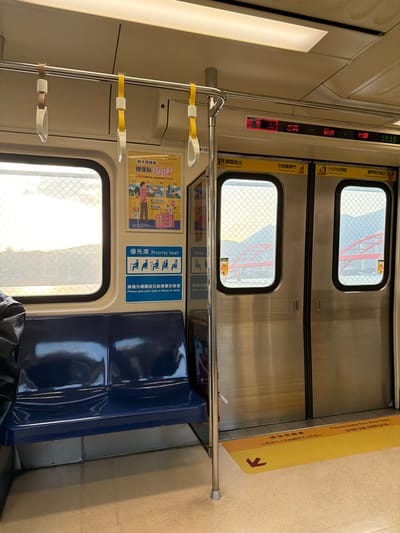The image presents the interior of a Taoyuan Airport MRT train, strongly indicating its operation within or near New Taipei City, Taiwan, given the visible station names "林口" (Linkou) and "竹圍" (Zhuwei) on the overhead digital display. The train car features a clean, light beige ceiling and walls, illuminated by overhead fluorescent light panels. A silver horizontal grab bar runs along the ceiling, from which three white and yellow hanging strap handles are visible. Below, two dark blue, glossy priority seats are situated on the left, with a dark-colored jacket or bag partially visible on the leftmost seat. A silver vertical support pole stands adjacent to the seats and train doors. On the right, two stainless steel train doors are closed, each featuring a small, rounded-rectangle window with a fine mesh pattern. Through these windows, a bright outdoor landscape is visible, including a prominent red arch bridge structure and hazy mountains in the distance. The digital display above the doors shows "林口" (Linkou), "竹圍" (Zhuwei), and the time "16:16". A yellow poster with Chinese text and illustrations, advertising "捷運點數" (MRT points), is affixed to the wall above a blue "優先座 Priority Seat" sign, which displays pictograms for elderly, pregnant, injured, and parent with child passengers. The floor is a light, uniform color, marked with a yellow floor decal near the doors, bearing a red arrow and multilingual text instructing to "Please keep this area clear." No individuals are directly visible within the frame.