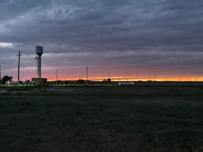 The image depicts a vast, open landscape at dusk. A prominent subject is a tall water tower with a metal ladder on its side, standing near a low-rise building with a brick facade. Several utility poles are scattered across the scene, with wires stretching between them, suggesting a rural or semi-urban setting. In the middle ground, a white car is visible on a road, its headlights illuminated, indicating movement. The foreground is a dark, grassy field.
The sky is dramatic, filled with heavy, dark gray clouds that are beginning to break apart, allowing a vibrant band of orange and pink light from the setting sun to illuminate the horizon. This transition from day to night, coupled with the cloud cover, creates a mood of quiet contemplation or perhaps a sense of approaching storm. The overall setting appears to be Öndiris, Kazakhstan, characterized by its flat terrain and the presence of infrastructure like the water tower and utility poles. There is no visible text in the image.