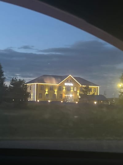 The image shows a two-story yellow building with a dark roof, illuminated by yellow lights around its perimeter and windows. The word "МОТЕЛЬ" (Motel) is spelled out in illuminated white letters on the front of the building. The building appears to be set back from a dark road, with trees and some other smaller structures visible in the foreground and to the sides. The sky is a dusky blue with some clouds, suggesting it is evening or early morning. The building is located in the city of Yamany, Russia.