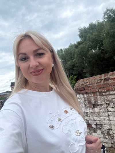 A woman with blonde hair and light skin is taking a selfie outdoors. She is wearing a white t-shirt with decorative embroidery and embellishments. She has on makeup, including lipstick and mascara, and is wearing a pearl earring. Behind her, on the right, is a weathered brick wall. In the background, to the left, there are trees and a cloudy sky. The location is identified as Verkhotor, Russia.