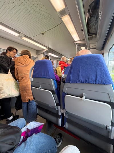The image shows the interior of a passenger train, likely in transit. Several passengers are visible, seated in rows of blue and gray seats with folding tables. An overhead compartment contains luggage, and overhead lights illuminate the scene. The presence of Cyrillic characters on a bag in the foreground suggests a location in Russia. There is no information in the image that indicates this image was taken in the city of Lyuban, Russia.