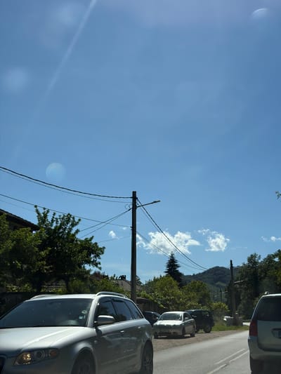 The image captures a view of a street in Beli Osam, Bulgaria, on a sunny day. The sky is a vibrant blue with scattered white clouds. Power lines are visible, strung between utility poles, and a street lamp is mounted on one of them. In the foreground, a silver station wagon is parked. Further down the street, other vehicles are present, including a silver sedan and a dark SUV. Trees and houses line the sides of the road, and in the background, lush green hills rise, indicating a natural, possibly rural or semi-rural setting. The overall impression is one of a typical village scene on a bright, clear day.
