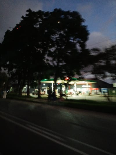 This blurry, low-light photograph captures a roadside scene at dusk or night in Marondera, Zimbabwe. The image is vertically oriented and appears to be taken from a moving perspective, resulting in significant motion blur.

On the left side, a large, dark tree dominates the foreground, silhouetted against a muted blue-grey sky with faint clouds. The tree's branches and foliage are indistinct due to the darkness and blur. On the right, a brightly illuminated commercial establishment, likely a gas station or convenience store, stands out against the dim surroundings. Its lights, both from within the building and from overhead canopies, cast a strong glow on the immediate area. Faint outlines of multiple fuel pumps are visible beneath the canopies. Several blurry shapes in front of the building suggest the presence of vehicles, possibly parked or pulling in.

A dark asphalt road with discernible white lane markings runs vertically along the far right edge of the frame. The overall atmosphere is dark, with artificial lights being the primary source of illumination. There are no clearly identifiable people. While some shapes on the building might be part of signage, no text is legible due to the blur and low resolution. A white "F" shape is faintly visible in one of the brighter areas, potentially part of a brand logo. The scene depicts typical evening activity along a commercial road.