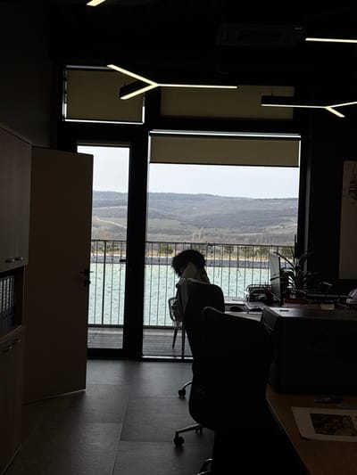 The image depicts an interior view of an unoccupied modern office space with an extensive exterior vista, situated in Зеленогорское сельское поселение, Ukraine. The indoor environment features a dark ceiling with two illuminated Y-shaped linear LED fixtures and an overhead air conditioning vent. Light-toned wooden cabinetry containing binders is positioned along the left wall. The floor consists of dark square tiles. A large glass facade, comprising a partially ajar door on the left and windows with lowered roller blinds on their upper sections, forms the rear boundary. Inside, several dark office chairs are observed; one positioned near the window is draped with a dark, textured material. A workstation adjacent to the window includes a laptop, a potted plant, and a desktop organizer. Papers with visual content are visible on a desk in the immediate foreground. The exterior view reveals a wooden deck bordered by a metal railing, overlooking a distinct turquoise-green body of water. Beyond the water, a distant landscape of undulating hills, characterized by sparse deciduous vegetation and visible patches of earth, extends under an overcast sky. No human presence or activity is observable within the frame.