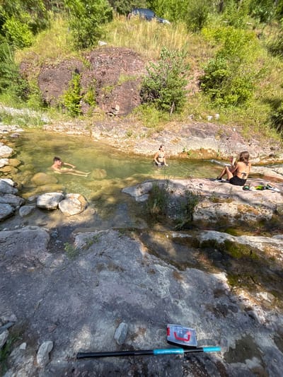 A man in yellow swim trunks is submerged in a clear, shallow natural pool of water surrounded by large rocks. He is floating on his back, facing the camera, with his arms extended. To the right, on the bank, a woman in a black bikini sits on a rock, facing away from the camera, holding a pink object. Another woman, also in a black bikini, stands in the water near her, facing the sitting woman. The environment is natural and outdoors, with a rocky slope covered in grass and trees in the background. A black pole with blue markings and a clear plastic container with red objects inside are on the rocks in the foreground. The scene appears to be a secluded spot within the Zgorigrad region of Bulgaria.