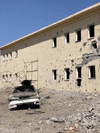 The scene depicts a damaged two-story building and a destroyed vehicle under a clear blue sky in Bar Mel, Afghanistan. The light beige building exhibits extensive damage, including numerous small, circular impact marks across its entire facade and larger sections of concrete and stucco blown out, particularly on the lower right side. All visible window openings are empty, lacking glass, with some frames intact and others damaged. In the foreground, a heavily damaged white sedan is centrally positioned. Its roof and upper structure are severely caved in and mangled. The front bumper is dislodged, the hood is crumpled, and the windshield is absent. A partially legible white license plate with black text is visible on the front of the vehicle. Behind the car, a rusted, bent metal framework, possibly remnants of scaffolding or a structural frame, stands between the vehicle and the building. The ground is entirely covered in debris, consisting of various sizes of rocks, concrete fragments, and scattered litter. No people are present.