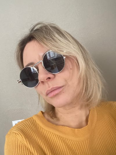 A woman poses, wearing round sunglasses with a silver frame. Her hair is blonde with darker roots. She's dressed in a mustard-yellow sweater with a ribbed texture. The background is a neutral, light-colored wall. The image is slightly tilted to the right. The angle is from a higher perspective, looking down at her face.