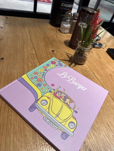 A high-angle shot captures a tabletop scene, featuring a book titled "Le Burger" with a graphic of a yellow Volkswagen Beetle on its cover, alongside a decorative glass vase containing plant life and some condiments. The setting is likely indoors, given the polished wooden surface and interior lighting. The location cannot be determined to be in Amstetten, Austria based on provided visual elements.