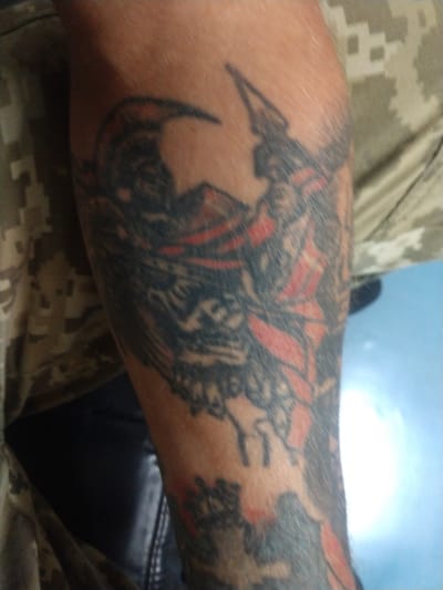 This is a close-up, waist-up shot of a person's forearm with a tattoo, taken outdoors in Vylok, Ukraine. The person is wearing a camouflage shirt. The tattoo appears to be a stylized depiction of a winged creature or warrior, with predominantly black and red ink. The background consists of a blurry, light blue sky with a bright, out-of-focus light source, possibly the sun. The image quality is slightly grainy and the focus is on the tattoo itself.