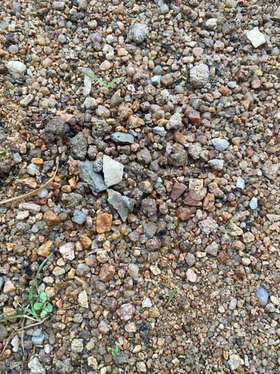The image is a close-up, overhead view of a textured surface composed of small, irregular stones and gravel. The gravel consists of a mix of colors, including shades of brown, gray, white, and some reddish-orange hues. The stones vary in size and shape, with some being smooth and rounded, while others are rough and angular. A few small, sparse green blades of grass are scattered across the gravel. There is also a single dried, straw-like stem lying horizontally in the lower portion of the image. The lighting appears to be natural, suggesting it might be daytime, and there are no strong shadows, possibly indicating overcast conditions or diffused sunlight. The setting is likely an outdoor area, such as a path, yard, or unpaved ground in Dumnga, Nigeria, given the location context. There is no discernible activity or event taking place; the image focuses solely on the static composition of the gravel. No text is visible in the image.