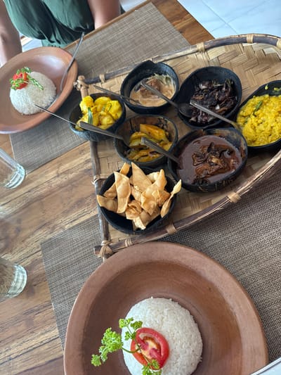 The image shows a spread of Sri Lankan cuisine. A woven tray holds six small, dark bowls containing various dishes: yellow curry, a dark-colored meat curry, a lighter creamy curry, pineapple curry, fried wonton strips, and a vegetable curry. Two terracotta bowls hold mounds of white rice, each topped with a tomato slice and parsley. The scene is likely set indoors, given the wooden table and glimpses of a person in a green shirt. The setting could be a restaurant or home in Polonnaruwa, Sri Lanka, a location known for its culinary offerings. Two drinking glasses are placed on the table.