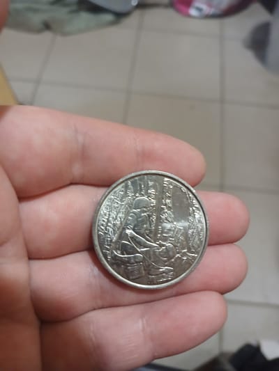 A close-up, slightly blurry image shows a person's hand holding a silver-colored coin. The coin features an embossed design depicting a figure, possibly a worker, in a wooded area. The background of the image is out of focus and appears to be an indoor setting with a tiled floor and a hint of other objects. The location in Chutove, Ukraine is not discernible from the image itself.