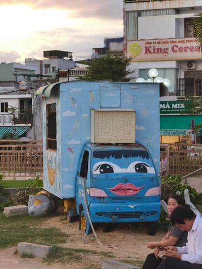 The image depicts a brightly painted blue truck with cartoonish eyes and lips, adorned with drawings of birds and clouds. The truck appears to be stationary, with one of its tires tilted outwards. Beside the truck, two individuals are seated on the ground, facing away from the camera. The man on the right wears a white collared shirt and is looking at his phone, while the woman next to him, wearing a striped shirt, looks towards the truck.
In the background, several buildings are visible. The closest building features a prominent sign with text in yellow and red, reading "King Cerea." Above this sign, another sign is partially visible with Vietnamese text. Further in the background, more buildings of varying heights and architectural styles are present, some of which appear to be residences or shops.
The scene is set outdoors, with a sandy ground area in the foreground where the truck and people are situated, and grass and vegetation to the left. A wooden fence runs along the middle ground. The sky above is filled with clouds, with hints of sunset colors. The overall impression is a street scene or an outdoor gathering area within a Vietnamese city, possibly An Nhơn, Vietnam.