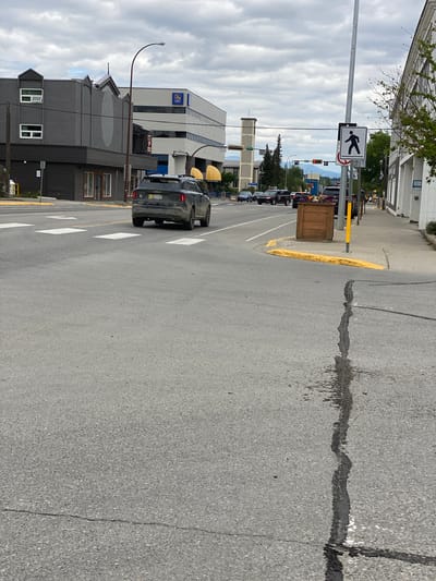 The image shows a view from a street in Whitehorse, Canada. A black Kia Sorento SUV is driving away from the camera, heading into an intersection. The SUV has a yellow license plate. In the background, there are several buildings, including a modern, multi-story building with windows and what appears to be a sign with "RBC" on it. A traffic light is visible, showing red. A pedestrian crossing sign is also present on a pole. The street is paved and marked with white crosswalk lines. There is a wooden planter box on the sidewalk. The sky is overcast. The image appears to be taken during the daytime.