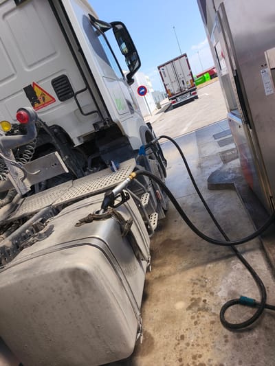 The image shows a white semi-trailer truck being refueled at a gas station on a bright, sunny day. The perspective is from a low angle, looking up at the truck's side and the fuel pump. A black fuel hose runs from the pump to the truck's large, dusty fuel tank, with the nozzle inserted into the tank opening. The ground is concrete, stained with dark marks, likely from oil or fuel spills.
In the background, another white truck with a trailer is visible further down the fueling lane. The sky is clear blue.
Notable details on the main truck include a yellow triangular warning sticker on the cab that reads "PELIGRO! ÁNGULO MUERTO" (DANGER! BLIND SPOT), depicting a person near a turning truck. A green logo, partially visible, appears to spell "bell" or "beli" on the side of the cab. The truck has prominent side mirrors.
The truck in the background has "SCHMITZ" written on its mudflap and a "Jost Group" logo on its trailer. A blue circular sign with a white truck icon, possibly indicating a traffic regulation, is also visible in the distance. The fuel pump to the right has some indistinct text and numbers, including "9-19", possibly indicating hours of operation. No people are visible in the scene.