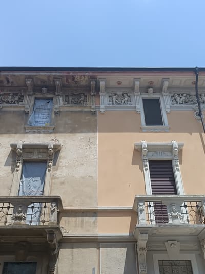The image shows the facade of a building in Casalmaggiore, Italy, divided into two sections. The left side appears to be undergoing renovation or is in disrepair, with weathered, peeling plaster and windows covered with protective sheeting. Ornate architectural details, including sculpted figures, are visible above the windows. The right side of the building is freshly painted in a pale peach color and features pristine white decorative elements around the windows and balconies. The contrast between the two sides suggests a restoration project. The sky above is clear and blue. There are no people visible in the image.