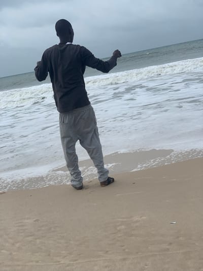 An individual stands facing the Atlantic Ocean on a coastal beach in Ibeju, Nigeria, under an overcast sky. The subject, an adult male viewed from the back, wears a dark long-sleeved shirt, light grey sweatpants, and dark-colored sandals. His arms are bent at the elbows, with hands clenched into fists and held slightly outward. White-capped waves break and recede onto the wet, light brown sand at the shoreline. The foreground consists of damp, rippled sand. A small, white, rectangular object is visible on the sand in the lower right portion of the frame.