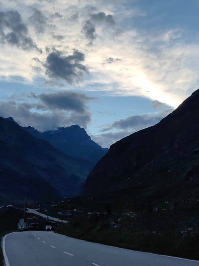 The image shows a scenic view of a mountain road in the evening, likely in the region of Damphug, India, given the context. The road, paved with asphalt and marked with white lines, curves through a valley flanked by steep, dark mountains. The sky above is a dramatic canvas of clouds, with patches of bright light breaking through, suggesting either sunrise or sunset. A scattering of small buildings and structures, possibly a village, are visible in the lower part of the valley, nestled against the mountainside. A few vehicles, including what appears to be a truck and a smaller van, are present on the road, their headlights faintly illuminated. The overall atmosphere is serene and majestic, capturing the grandeur of the Himalayan landscape at dusk.
