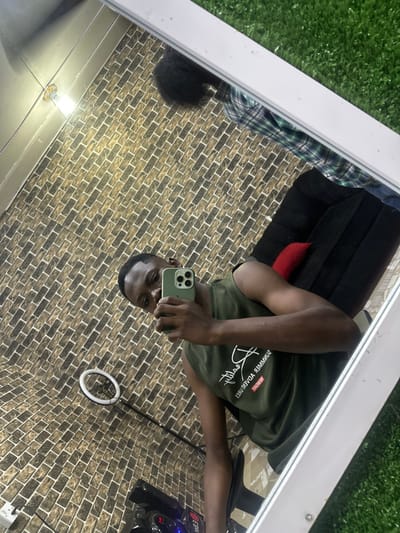 A person takes a mirror selfie, holding a light green iPhone. They wear an olive green t-shirt with white writing. The background appears to be a room with brick-patterned walls. A black chair is visible in the reflection along with a motorcycle. A ring light is set up near the subject. Grass-like material is partially visible near the top left, next to a plaid shirt and dark pants, which suggests the photo was taken inside a shop.