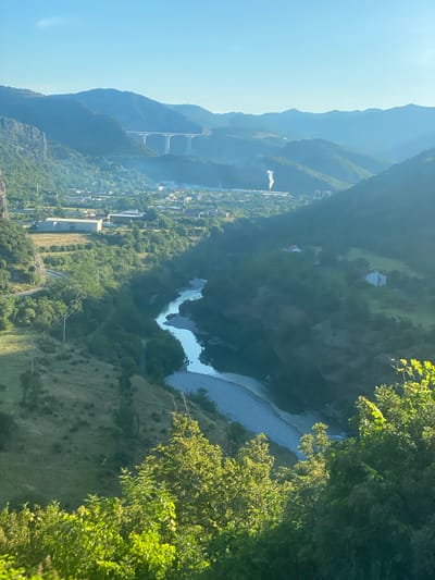 This is an elevated, wide-angle shot of a valley with a river flowing through it, located in the city of Donje Mrke, Montenegro. The river, reflecting the bright blue sky, curves through the valley floor. The surrounding hills are densely covered with green trees, with some areas of dry grass and rocky outcrops visible. In the middle ground, a small town or village is nestled within the valley, with various buildings and structures. A large, modern bridge with multiple pillars spans a distant section of the valley, and further in the distance, a plume of white smoke rises from what appears to be an industrial facility. The sky is clear and bright blue, with the sun casting a warm glow on the landscape, suggesting it is early morning or late afternoon. The foreground is dominated by the lush green foliage of trees and bushes, framing the view of the valley.