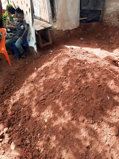 The image depicts a semi-sheltered outdoor area in Oko Town, Nigeria, characterized by reddish-brown laterite soil. A large mound of this soil, consistent with recent excavation, occupies the central and foreground. Dappled shadows are cast across the soil surface. To the left, a young male, approximately 8-12 years old, sits on a blue plastic chair, wearing a dark polo shirt and blue jeans, with flip-flops on his feet. Behind him, partially obscured, are at least two adult females seated on chairs, dressed in patterned clothing and one with a headscarf. An orange plastic chair is visible to the immediate left. A white perforated plastic or fabric sheet serves as a vertical screen, affixed to a pole, separating the seating area from the background. In the background, behind the screen, a makeshift wooden table is present, along with corrugated metal sheeting, dark tarpaulin, and another light-colored fabric or plastic sheet forming a rudimentary wall structure. The ground beneath these background elements is also exposed reddish-brown earth.