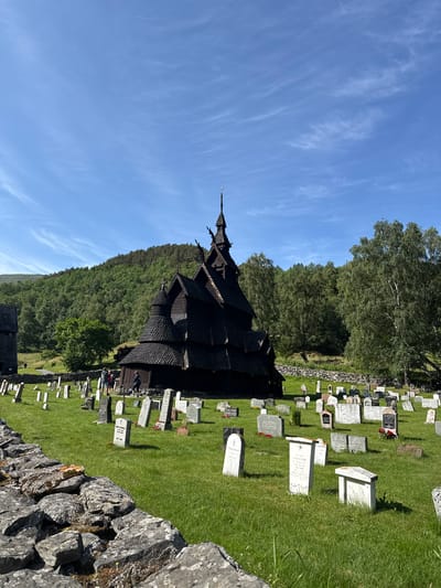 The image depicts the Borgund Stave Church in Borgund, Norway, a historic wooden church dating back to the late 12th century. The church, with its distinctive dark wood and steep, tiered roofs, is the central subject of the scene. It is surrounded by a graveyard filled with numerous gravestones of varying sizes and shapes, most of which are made of light-colored stone. The ground is covered in lush green grass. In the background, a dense forest of green trees covers a rolling hillside under a clear blue sky with sparse, wispy white clouds. A low stone wall runs across the foreground, partially obscuring the view of some of the gravestones. Two individuals are visible near the entrance of the church, and a few other people are scattered throughout the graveyard. The overall atmosphere is peaceful and serene, suggesting a historical and commemorative site on a pleasant day.