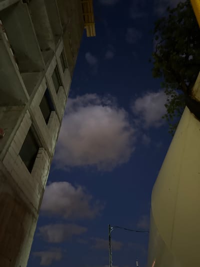 The image depicts an urban construction site at night, with a deep blue sky and scattered clouds. On the left, a partially constructed building made of concrete and white blocks is visible, with dark, empty window openings. Yellow scaffolding is attached to the top of the building. To the right, a large, curved, cream-colored structure, possibly a silo or part of another building, dominates the foreground. A dark green tree with dense foliage is seen against the sky to the upper right. Electrical wires and a street lamp pole are visible in the lower center. The overall impression is one of ongoing development in a city at dusk or night. The scene is consistent with a construction site in the city of Moscow, Russia.